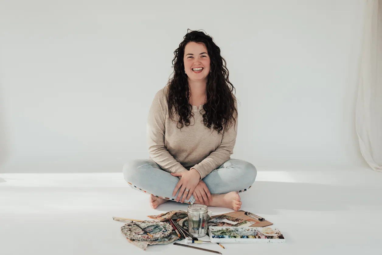 Alyssa Whetstone '19 is wearing a tan long-sleeved shirt and white washed jeans and is sitting in front of watercolor painting supplies in a photography studio