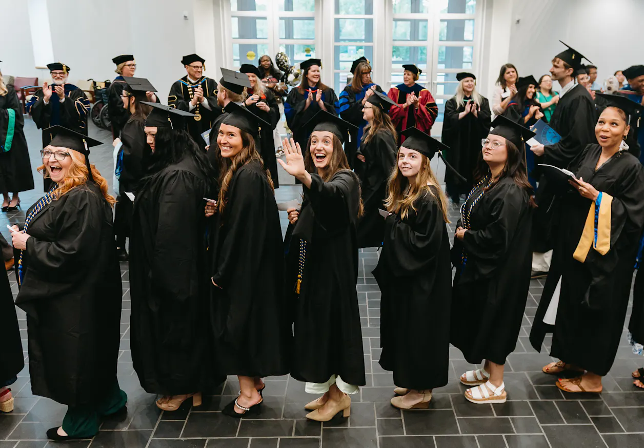 Graduates gather before commencement as one student waves to the camera.