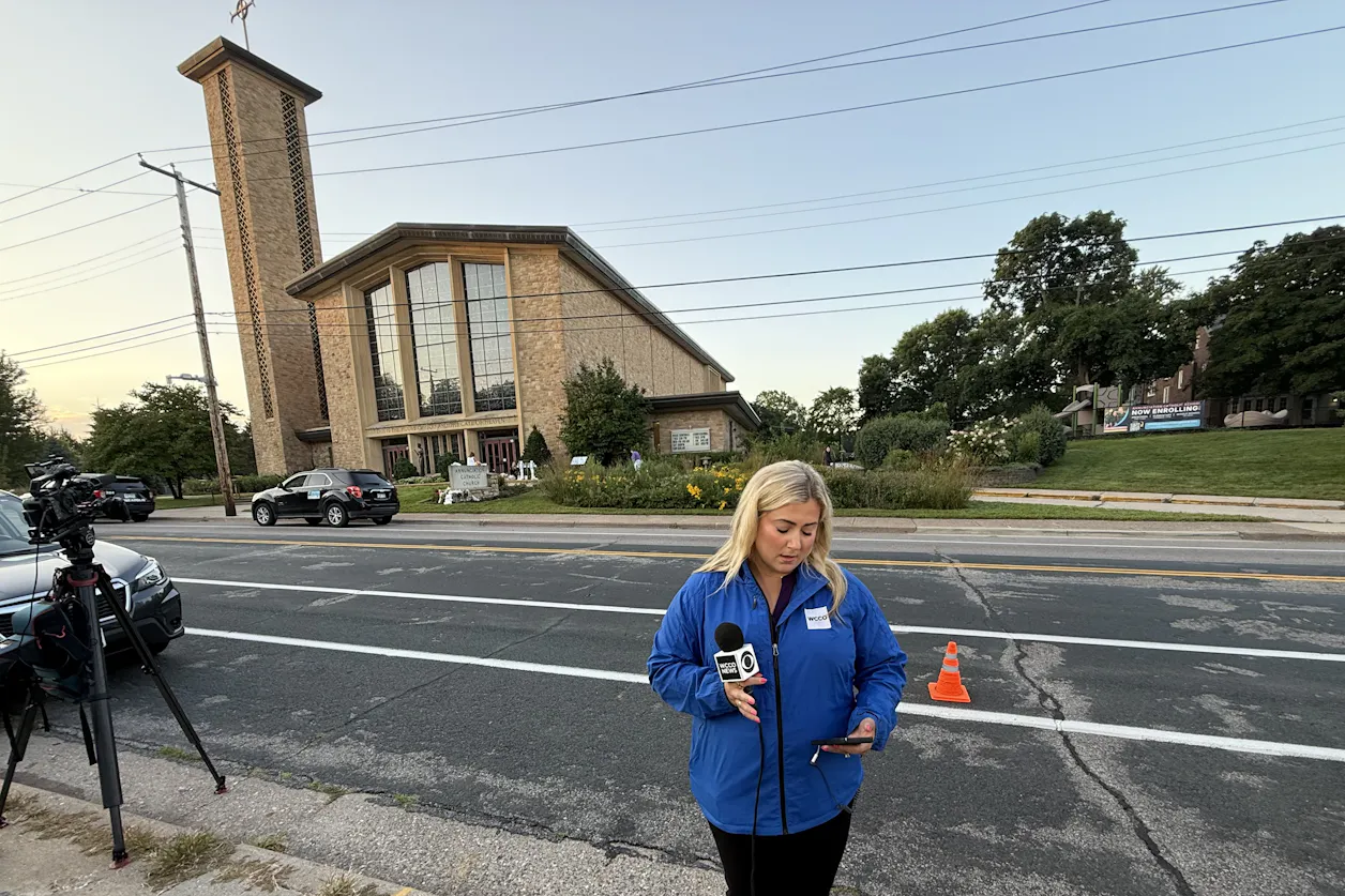 Beret Leone is wearing a blue WCCO windbreaker with a microphone in her hand while standing in front of a church-Annunciation Catholic School. 