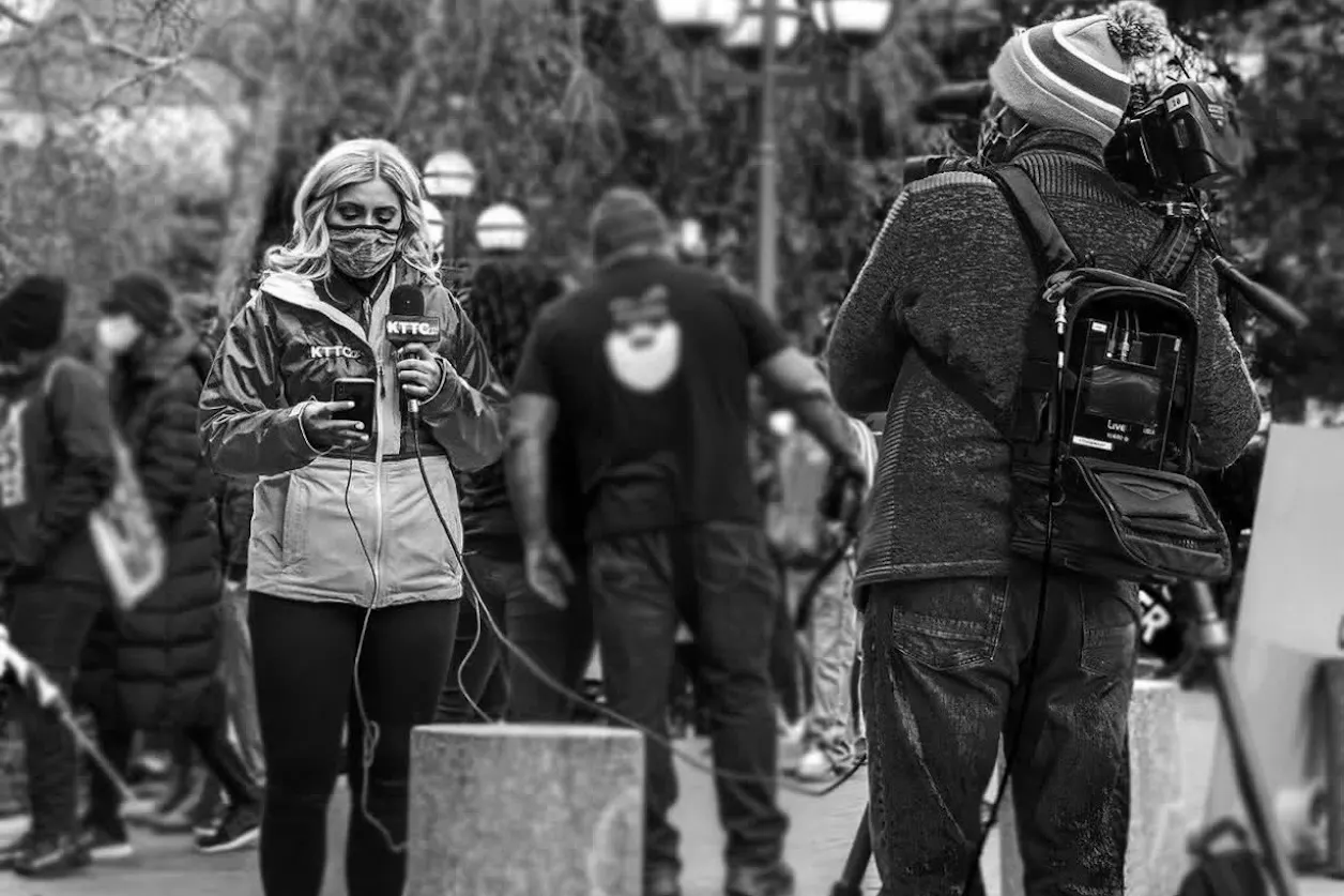 Beret Leone stands connected by wires to her camera man. She is wearing a mask and the photo is in black and white. 