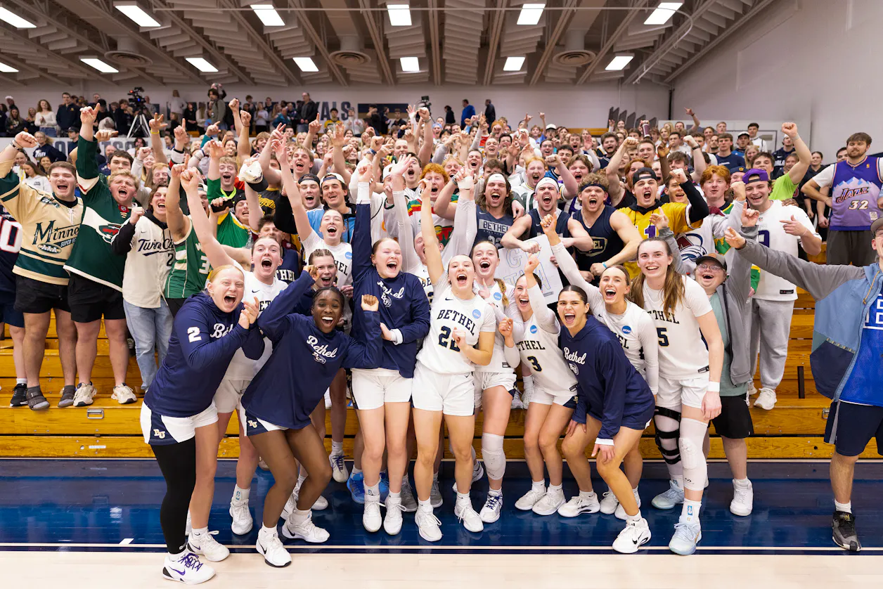 A group of students gather around the women's basketball team on the court to cheer and smile at the camera.