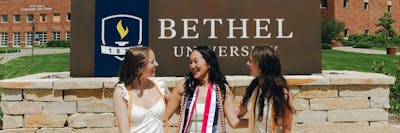 Three graduates in white dresses and honor cords smile together in front of the Bethel University sign.