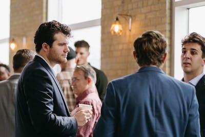 Men in suits converse during a professional networking event in a warmly lit, brick-walled space.