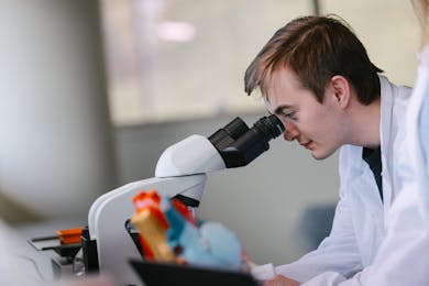 Student in a lab coat looks through a microscope during a hands-on science class at Bethel University.