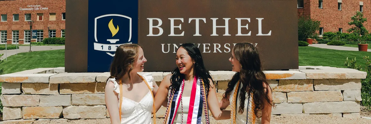 Three graduates in white dresses and honor cords smile together in front of the Bethel University sign.