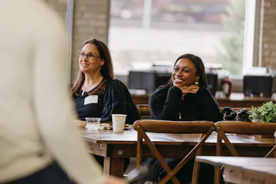 Two women seated at a wooden table smile and listen during a casual professional gathering.