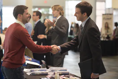 Employer and student shake hands at a career fair booth surrounded by conversation and displays.