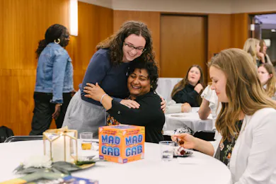 Smiling woman hugs a seated friend during a joyful gathering with games and dessert.