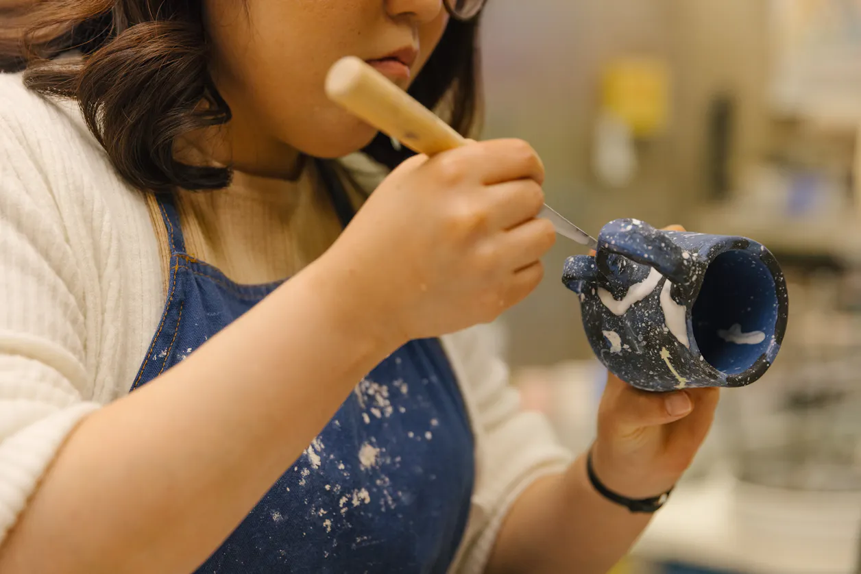 Art student meticulously carving intricate designs into a ceramic mug.