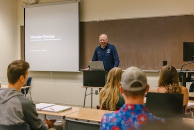 A Bethel University student presenting in front of the class, with a slide titled 'Missional Theology' displayed on the screen behind him.