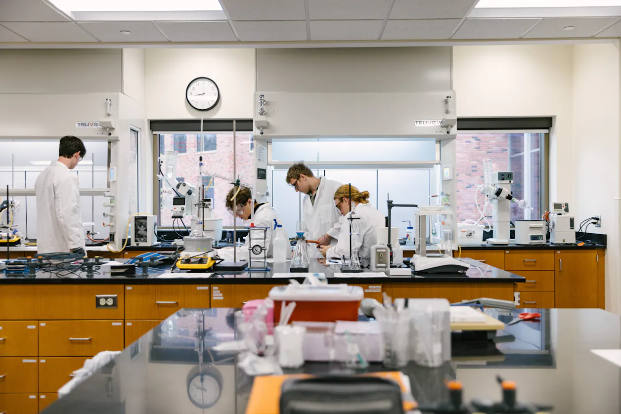 Four students in white lab coats work together at a lab bench with various equipment.