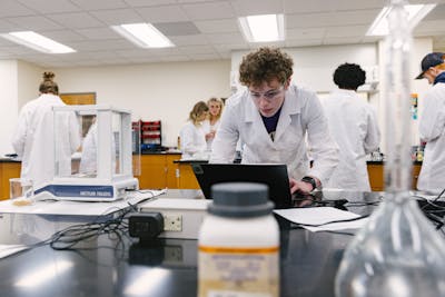 A student in a lab coat concentrates on a laptop, surrounded by lab equipment.