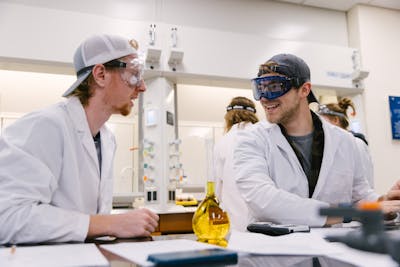 Two students in lab coats share a light moment while conducting an experiment.