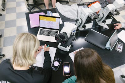 Students collaborate on a biology project, using laptops and microscopes.