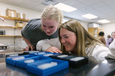 Two smiling students bend down to peer at samples in a science laboratory classroom. 