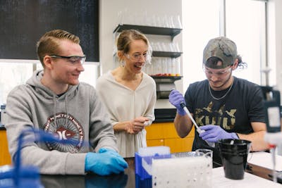 Students and a professor in a lab, engaged in an experiment with pipettes and safety goggles.