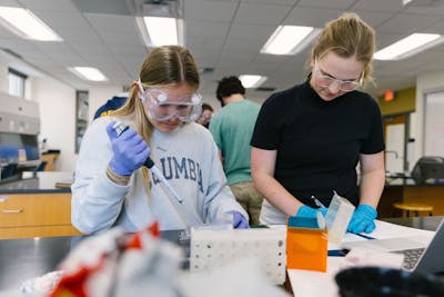 Two students in lab coats and goggles, carefully conducting an experiment together.