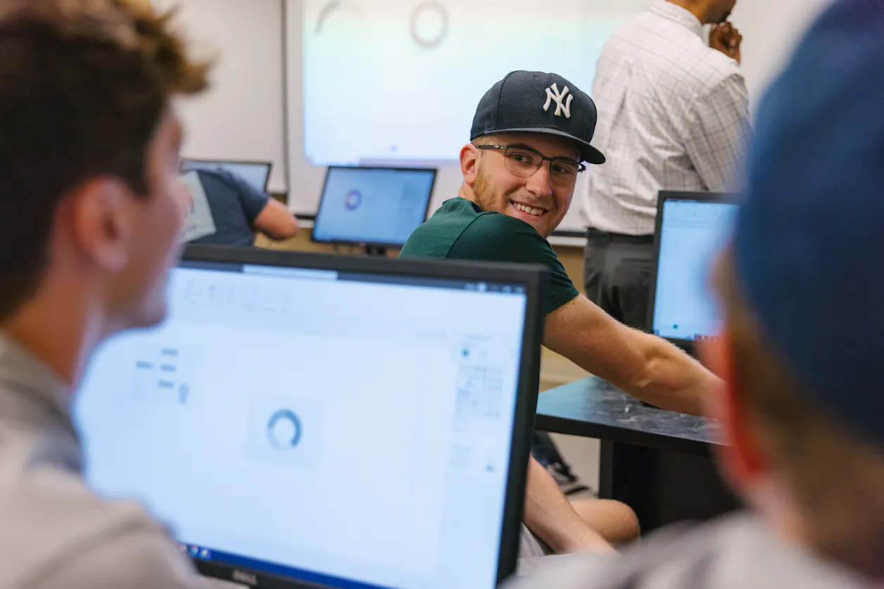 Students in a computer lab discussing a project, with one student smiling back at a student in the row behind.