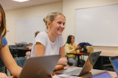 A Bethel University student smiling and working on a laptop during a class, with classmates in the background.