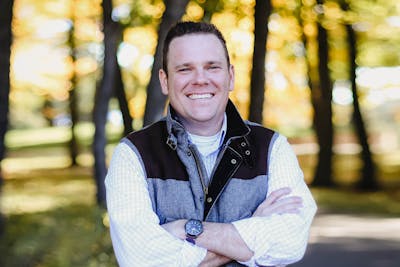 A man smiles in front of a wooded background.