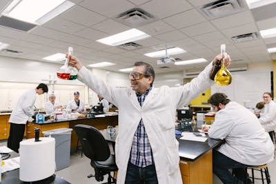 A professor demonstrates a science experiment to students, holding up flasks with colored liquids.