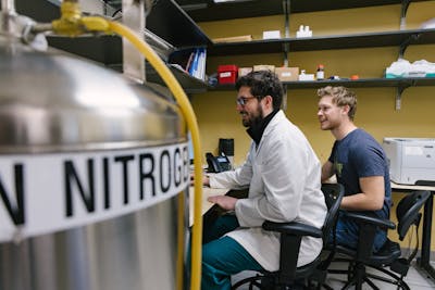 Two students work at a computer in a lab, with a liquid nitrogen tank visible.