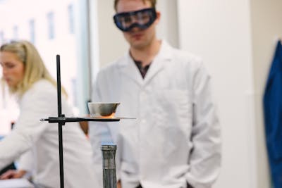 A student in safety goggles watches a flame under a beaker during a chemistry experiment.