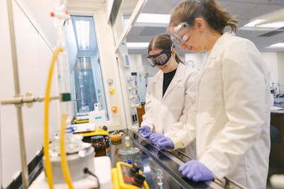 Two students in lab coats and goggles conduct an experiment in a chemistry lab.