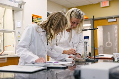 Two students in lab coats collaborate on a chemistry experiment, smiling and taking notes.