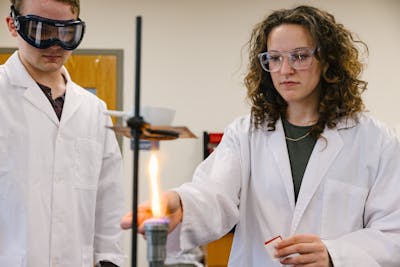 Two students in lab coats and goggles perform a flame test in a chemistry lab.