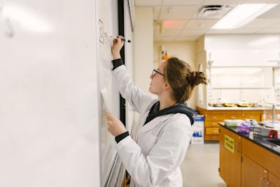 Student writing chemical equations on a whiteboard in a lab.