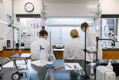 Three students in lab coats working together in a bright chemistry lab.