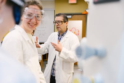 A professor in a white lab coat explains a concept to a smiling student.