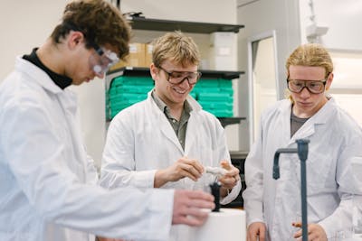 Three students in lab coats and goggles working with equipment. 