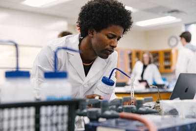 A Bethel University student focusing on a lab experiment, surrounded by lab equipment and wearing a lab coat.