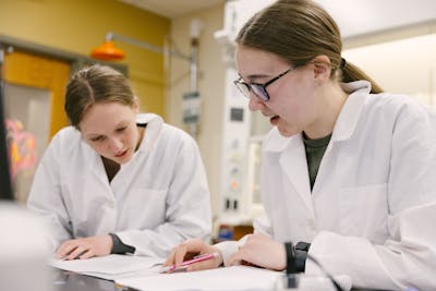 Two Bethel University students in lab coats working on a project together, looking at documents on the table.