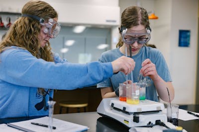 Two students in goggles and lab coats, working together on a colorful experiment.