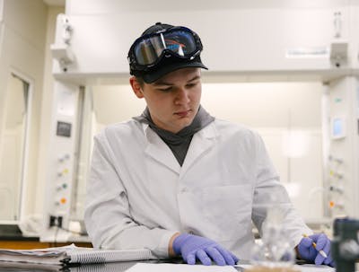 Student in a lab coat and safety goggles writing notes at a laboratory workstation.