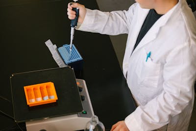 Student in a lab coat using a pipette to transfer liquid into a blue microplate in a chemistry lab.