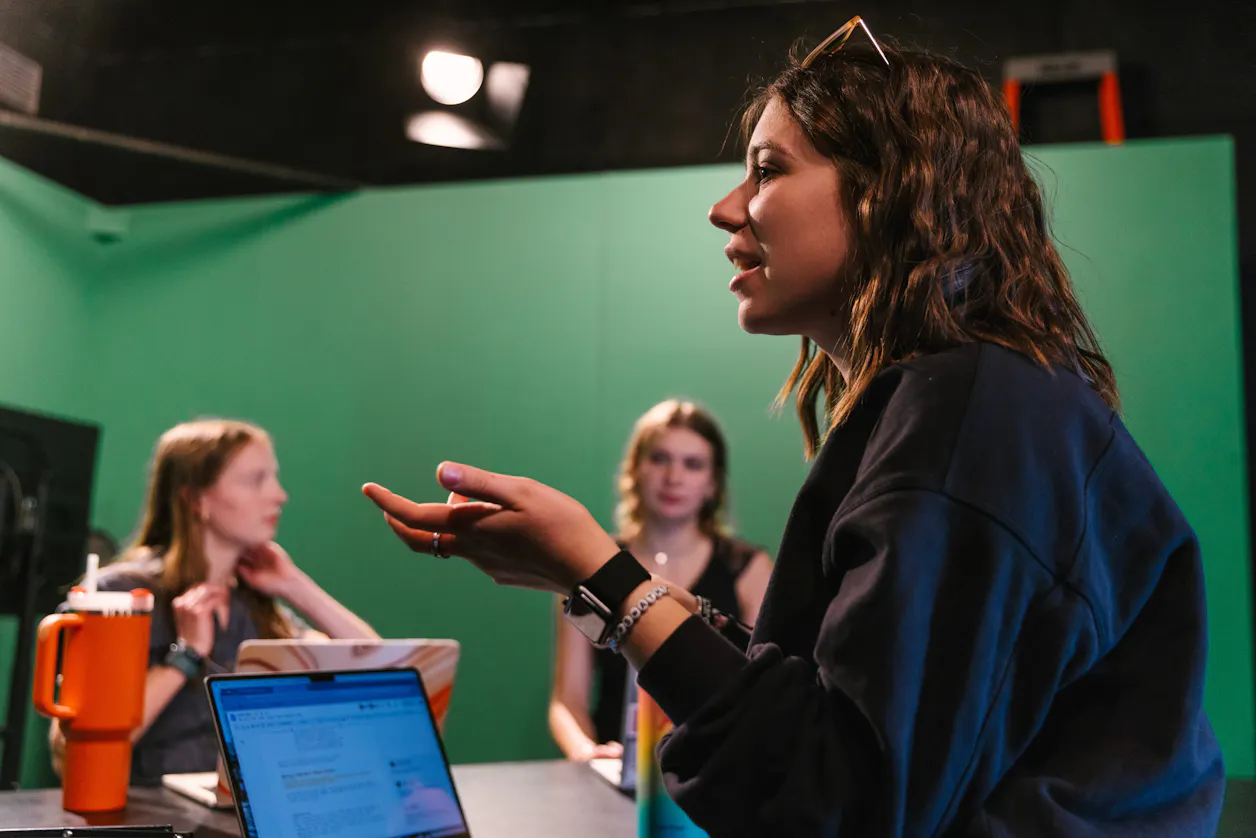 A student presenting in a green screen studio, others listen attentively with laptops open. 