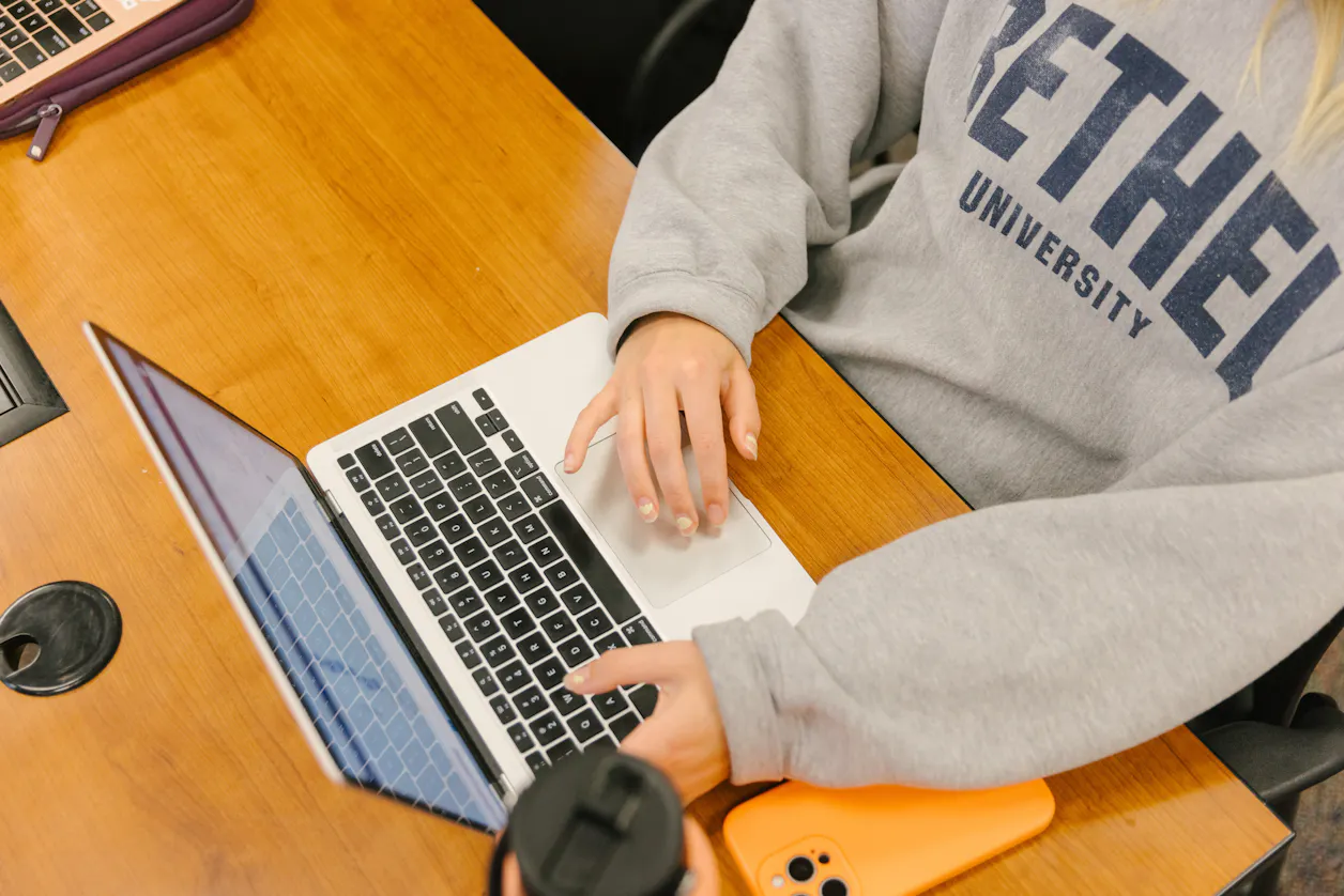 A close-up of a Bethel University student wearing a sweatshirt, working on a laptop in a classroom. 