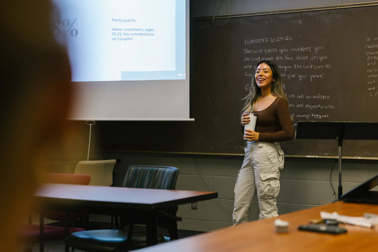 A student presenting research in front of a class, standing near a projection screen. 