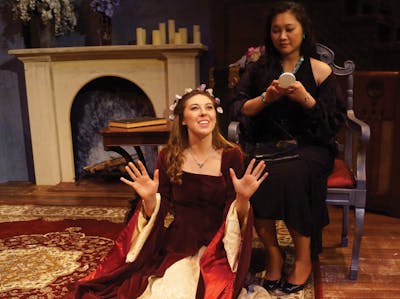 Two Bethel University students in a theatrical scene, one dressed in a medieval gown and the other holding a mirror.