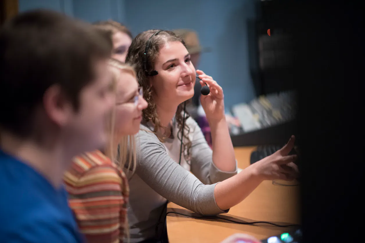 Bethel University students working together in a media production room, one wearing a headset and speaking into a microphone. 