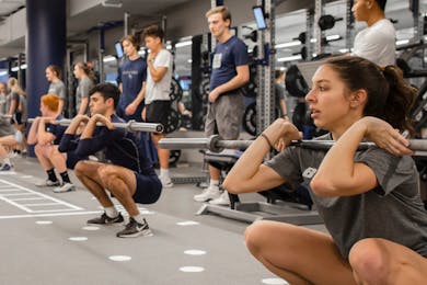 Students performing squats with barbells in a gym, engaged in strength training.