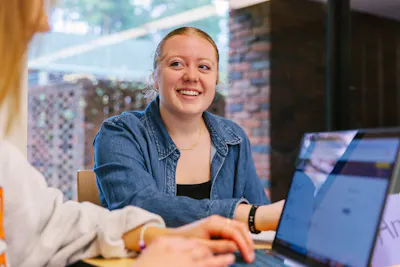 Student smiles while collaborating with a classmate beside a laptop.