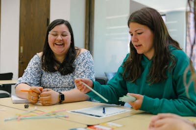 Two students laugh and discuss while working with colorful crafts at a table in a Bethel University classroom.