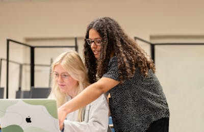 A teacher leans forward to share a laptop screen with student, engaging in an explanation during a class at Bethel University.