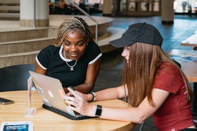 Two students smile while working together on a laptop in a bright campus space.