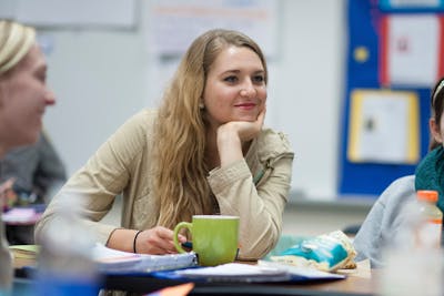 Student smiling with chin in hand during class, surrounded by notebooks, snacks, and a coffee mug.
